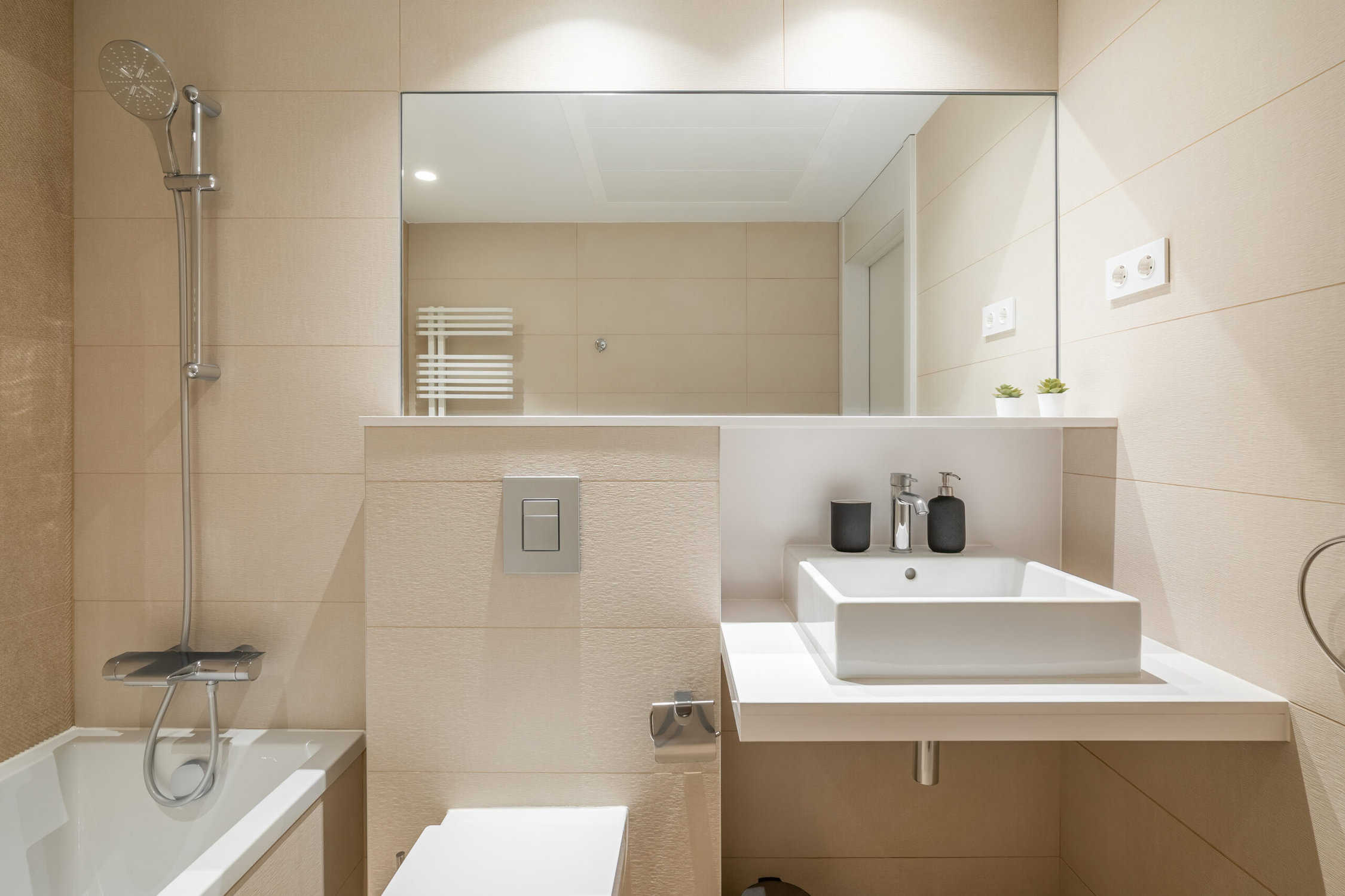 Cozy bathroom in modern design with huge mirror on the wall and bright lighting. Walls are in beige tiles and white sanitary ware. Room for taking morning shower and relaxing evening baths