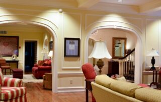 Image of a living room with some couches, chairs, lamps, and a staircase. The walls are painted a light yellow color with white trimmed accents and the ceiling is painted white with recessed lighting and exposed cross beams.